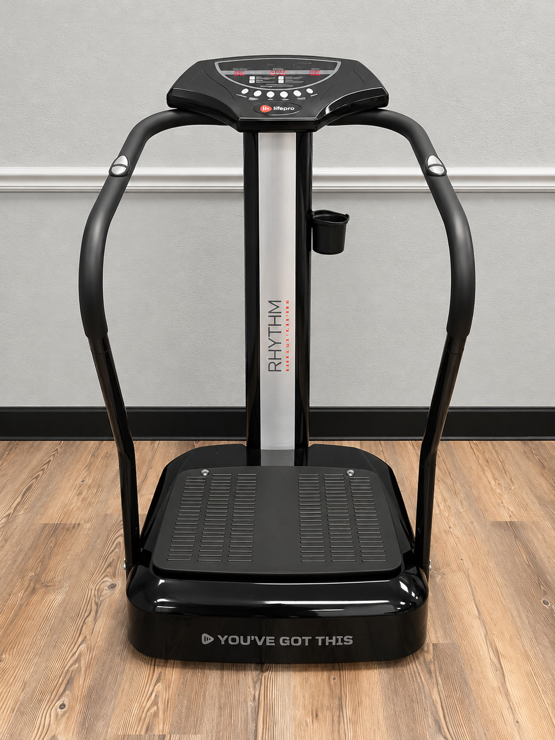 The vibration plate: a black upright platform machine with curved handlebars and a control panel, standing on the studio's wood floor.